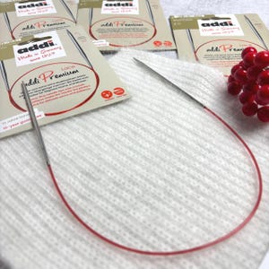 May include: A close-up of Addi Premium circular knitting needles with a red cable. The needles are silver and are packaged with beige and red cardboard. The packaging includes the text "Made in Germany Since 1829".