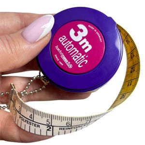 Puede incluir: Una cinta métrica automática de 3 m de color morado y rosa con una cinta amarilla y blanca. La cinta está marcada con números y la palabra "POLYESTER". El texto "MADE IN GERMANY" es visible. La cinta métrica se sostiene en una mano.