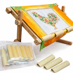 May include: A wooden embroidery stand with a colorful rooster design. The stand holds a fabric with a rooster embroidered in red, green, and yellow. Additional accessories include plastic scroll rods and a set of plastic clips.