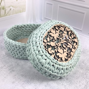 Könnte beinhalten: Ein hellgrüner, ovaler Häkelkorb mit einem Holzdeckel mit floralem Ausschnitt. Der Korb ist offen und zeigt einen hellfarbigen Innenraum. Der Deckel bedeckt den Korb teilweise.