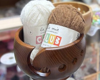 May include: A dark brown wooden yarn bowl with a spiral cutout and circular holes, containing two skeins of La Mia Club yarn. One skein is white and the other is brown, each featuring a paper label with the text 100 GR - 155 MT.