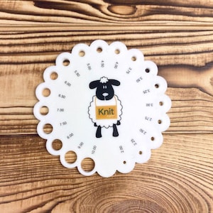 Op de afbeelding: Witte plastic breinaaldmaat met een zwart-witte schaapillustratie. De maat heeft een ronde vorm met gaten voor verschillende naaldmaten. Het schaap draagt een gele schort met het woord "Knit" erop. De maat heeft nummers van 2,00 tot 10,00.