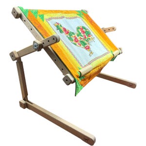 Puede incluir: Un soporte de bordado de madera con una tela colorida con un diseño de gallo. El soporte es ajustable y tiene un marco rectangular para sujetar la tela. La tela tiene un fondo amarillo y naranja.