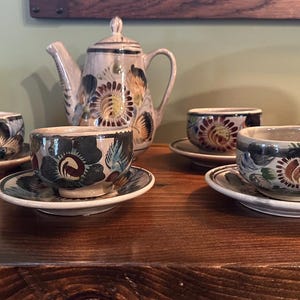 May include: A set of hand-painted ceramic teacups and teapot with floral designs in shades of blue, green, red, and yellow. The teapot has a rounded body and a spout on the side. The cups have a rounded shape and a small handle.