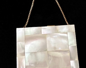 May include: A square, white clutch with a gold chain handle. The clutch is made of rectangular mother-of-pearl tiles. The gold chain handle is attached to the top of the clutch. The background is black.
