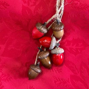 May include: A cluster of six decorative acorns, each with a unique color and texture, strung together with natural twine. The acorns feature a mix of red, brown, and tan hues, with textured caps. The background is a vibrant red fabric.