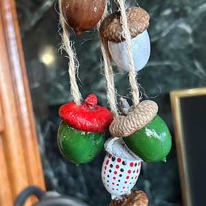 May include: A collection of painted acorn ornaments hanging from twine. The acorns are painted in various colors, including brown, white, green, and red. Some are decorated with polka dots. The ornaments are likely for seasonal or holiday decor.