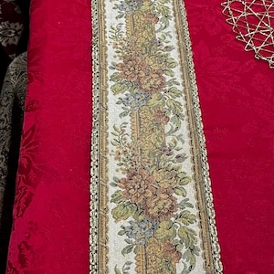 May include: A floral tapestry table runner with a central design of colorful flowers in shades of peach, green, and blue on a cream background. The runner is bordered with a gold trim and sits on a red tablecloth.