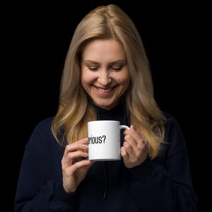 Op de afbeelding: Een vrouw glimlacht terwijl ze een witte koffiemok vasthoudt met de tekst "Serious?" in het zwart gedrukt.