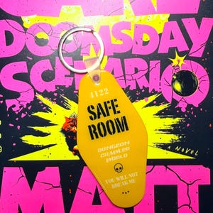 May include: Yellow Safe Room keychain with silver ring. The keychain has the number 4122 and the words "SAFE ROOM", "DUNGEON CRAWLER WORLD", and "YOU WILL NOT BREAK ME". The background is a book cover with pink and yellow text.