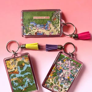 Puede incluir: Tres llaveros rectangulares de acr&iacute;lico con inserciones de mapas de parques tem&aacute;ticos a color. Un llavero tiene una borla rosa intenso, mientras que los otros dos presentan una borla combinada amarilla y morada. Cada uno incluye una argolla.