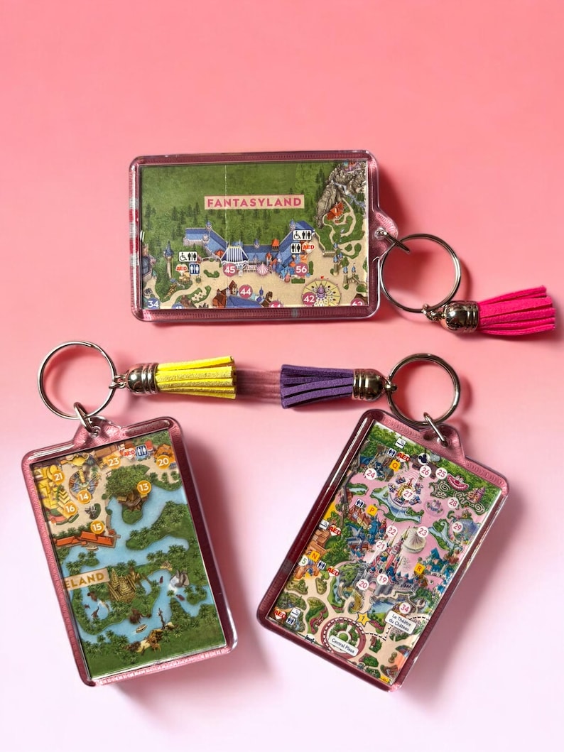 Puede incluir: Tres llaveros rectangulares de acr&iacute;lico con inserciones de mapas de parques tem&aacute;ticos a color. Un llavero tiene una borla rosa intenso, mientras que los otros dos presentan una borla combinada amarilla y morada. Cada uno incluye una argolla.
