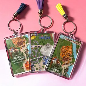 Puede incluir: Tres llaveros rectangulares de acr&iacute;lico con dise&ntilde;os de mapas de parques tem&aacute;ticos. Los llaveros de la izquierda y derecha muestran Tiana's Bayou Adventure, mientras que el central muestra Tron Lightcycle/Run. Cada llavero incluye una anilla de metal y una borla colorida.