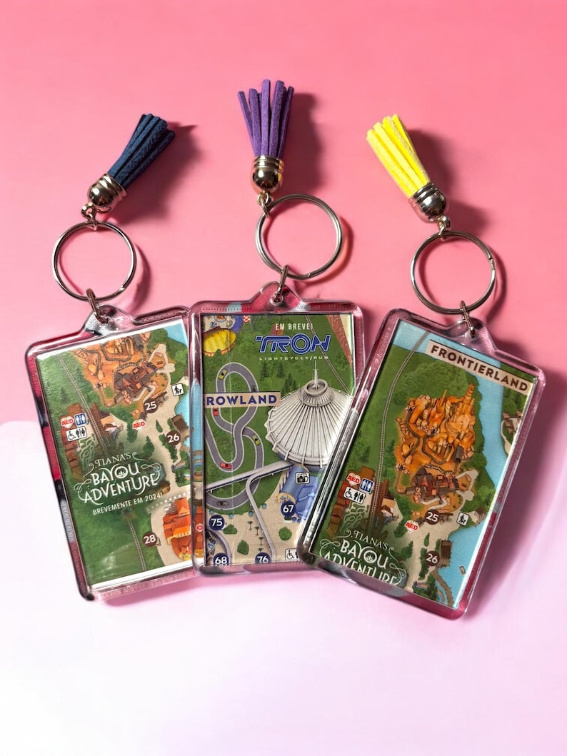 Puede incluir: Tres llaveros rectangulares de acr&iacute;lico con dise&ntilde;os de mapas de parques tem&aacute;ticos. Los llaveros de la izquierda y derecha muestran Tiana's Bayou Adventure, mientras que el central muestra Tron Lightcycle/Run. Cada llavero incluye una anilla de metal y una borla colorida.