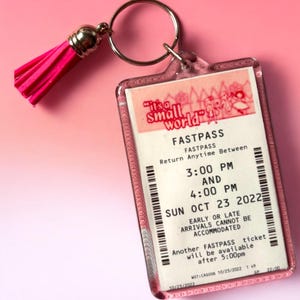 Puede incluir: Un llavero rectangular de acr&iacute;lico que presenta una r&eacute;plica de un boleto Fastpass de It's a Small World con texto negro sobre fondo blanco, incluyendo un anillo de metal plateado y una borla rosa brillante unida a la carcasa.