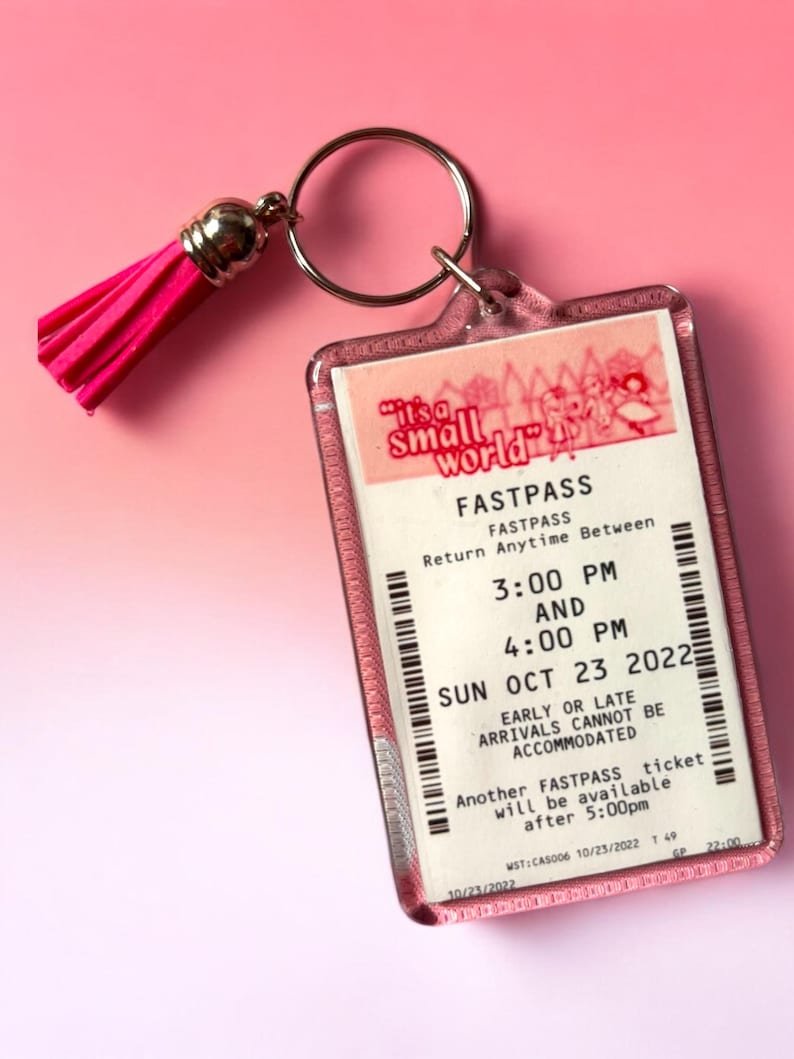 Puede incluir: Un llavero rectangular de acr&iacute;lico que presenta una r&eacute;plica de un boleto Fastpass de It's a Small World con texto negro sobre fondo blanco, incluyendo un anillo de metal plateado y una borla rosa brillante unida a la carcasa.
