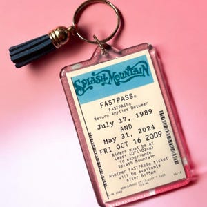 Puede incluir: Un llavero rectangular de acr&iacute;lico con una r&eacute;plica de un boleto Fastpass de Splash Mountain con las fechas 17 de julio de 1989, 31 de mayo de 2024 y 16 de octubre de 2009, incluyendo un requisito de altura de 102 cm, unido a una argolla dorada con borla azul.