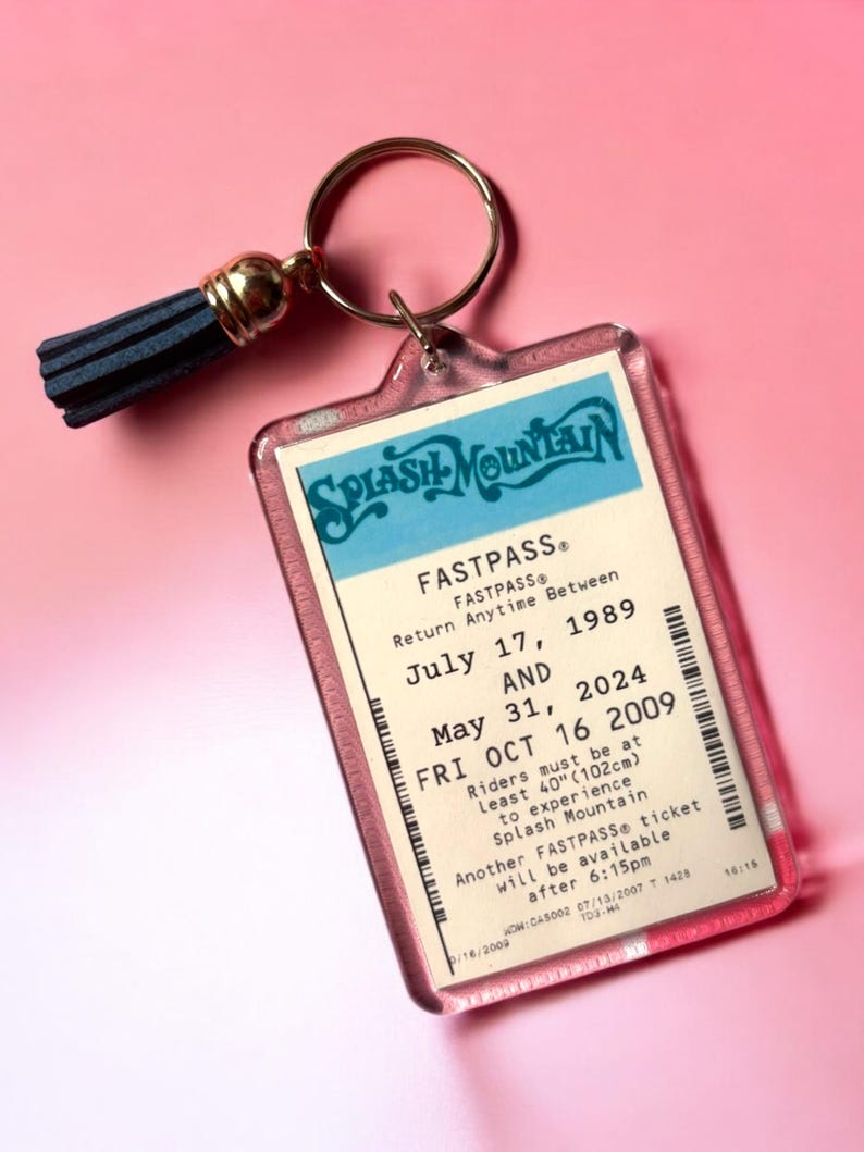 Puede incluir: Un llavero rectangular de acr&iacute;lico con una r&eacute;plica de un boleto Fastpass de Splash Mountain con las fechas 17 de julio de 1989, 31 de mayo de 2024 y 16 de octubre de 2009, incluyendo un requisito de altura de 102 cm, unido a una argolla dorada con borla azul.