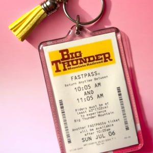 Puede incluir: Un llavero rectangular de acr&iacute;lico con una r&eacute;plica de un boleto FASTPASS de Big Thunder Mountain Railroad. El boleto incluye detalles de texto, un encabezado amarillo y un c&oacute;digo de barras. Una borla amarilla brillante est&aacute; unida al anillo de metal plateado.