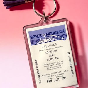 Puede incluir: Un llavero rectangular de acr&iacute;lico con una r&eacute;plica de un boleto Fastpass de Space Mountain con texto en morado, azul y blanco. El boleto incluye el texto Space Mountain, Fastpass y 10:50 AM a 11:05 AM. Una borla morada est&aacute; unida al anillo de metal.
