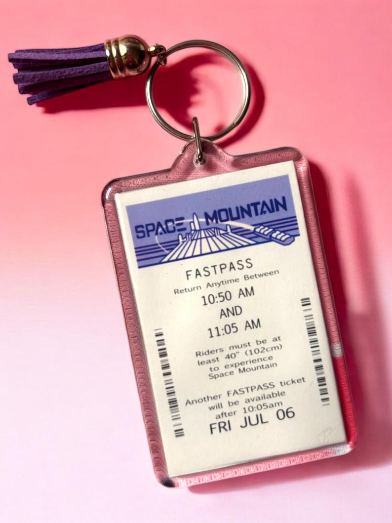 Puede incluir: Un llavero rectangular de acr&iacute;lico con una r&eacute;plica de un boleto Fastpass de Space Mountain con texto en morado, azul y blanco. El boleto incluye el texto Space Mountain, Fastpass y 10:50 AM a 11:05 AM. Una borla morada est&aacute; unida al anillo de metal.