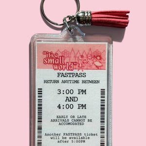 May include: A clear acrylic keychain featuring a rectangular insert designed as an It's a Small World Fastpass ticket. The ticket includes text, a barcode, and a pink tassel attached to a silver metal ring. The design is printed in black and pink.