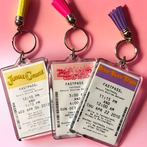 Puede incluir: Tres llaveros rectangulares de acr&iacute;lico con r&eacute;plicas de boletos Fastpass de parques tem&aacute;ticos para Jungle Cruise, It's a Small World y Peter Pan's Flight. Cada llavero incluye una anilla de metal plateado y una borla de ante en amarillo, rosa o morado.
