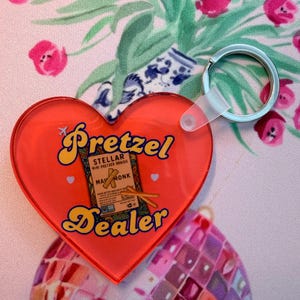 Puede incluir: Un llavero de acrílico rojo en forma de corazón con el texto "Pretzel Dealer" en una fuente retro amarilla. En el centro, un gráfico muestra una bolsa etiquetada como "Stellar Mini Pretzel Braids" de "May Monk" con dos pretzels. También se ven un pequeño icono de avión y dos corazones blancos.
