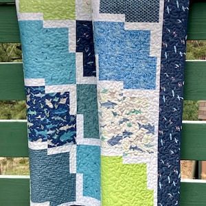 May include: A blue and green patchwork quilt with a white border. The quilt features a variety of ocean-themed prints, including sharks, fish, and waves.