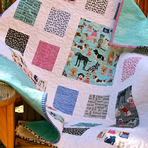 May include: A white quilt with a patchwork design featuring various dog and cat prints. The quilt is made up of squares with different patterns, including paw prints, stripes, and polka dots. The quilt is folded and draped over a wooden chair.