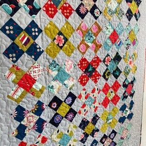 May include: A handmade quilt with a gray background and a repeating pattern of colorful, cross-shaped blocks. The blocks feature floral and geometric patterns in red, blue, yellow, and pink. The quilt is machine quilted with a wavy pattern.