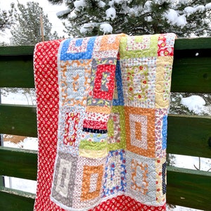 May include: A colorful patchwork quilt with a red, white, and blue floral pattern. The quilt is hanging on a green wooden fence against a snowy background.