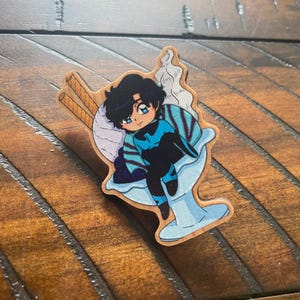 May include: A wooden pin featuring a cartoon character with blue hair and a blue and black outfit sitting in a blue and white ice cream sundae.