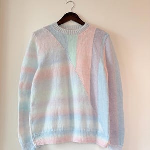 May include: A pastel-coloured jumper with a round neckline and long sleeves. The jumper features a colour-blocked design with sections of pink, blue, and green. The jumper is hanging on a wooden hanger against a white wall.
