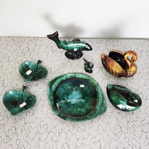 Puede incluir: Una colección de piezas decorativas de cerámica vintage. Incluye una figura de pez verde, un recipiente en forma de cisne, platos en forma de hoja, una pequeña figura de conejo y un plato grande y redondo. Las piezas tienen un acabado brillante con tonos verdes, marrones y dorados.