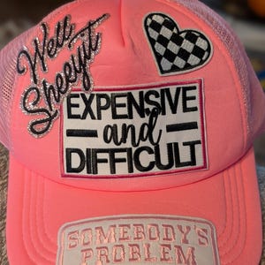 Peut inclure: Casquette de camionneur rose vif avec un dos en filet et une visière incurvée. La casquette présente plusieurs écussons brodés, dont un cœur à carreaux, la phrase "Well Sheeeyt" en paillettes argentées, et les mots "EXPENSIVE and DIFFICULT" et "SOMEBODY'S PROBLEM".