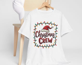 May include: White t-shirt with a Christmas Crew design. The design features a Santa hat, the words "Christmas Crew" in red, and a border of colorful Christmas lights. A white hat rests on a wooden chair.