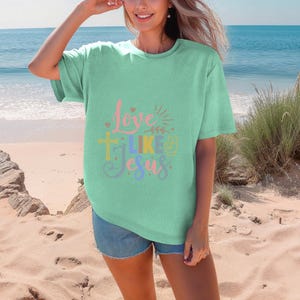 May include: A woman wearing a light green t-shirt with the text "Love Like Jesus" in colorful letters. The shirt has a cross and a peace sign graphic. She is standing on a sandy beach.