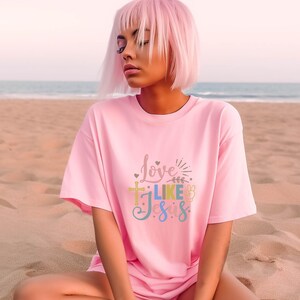 May include: A pink t-shirt with the text "Love Like Jesus" in colorful letters. The shirt features a cross, a peace sign, and hearts.