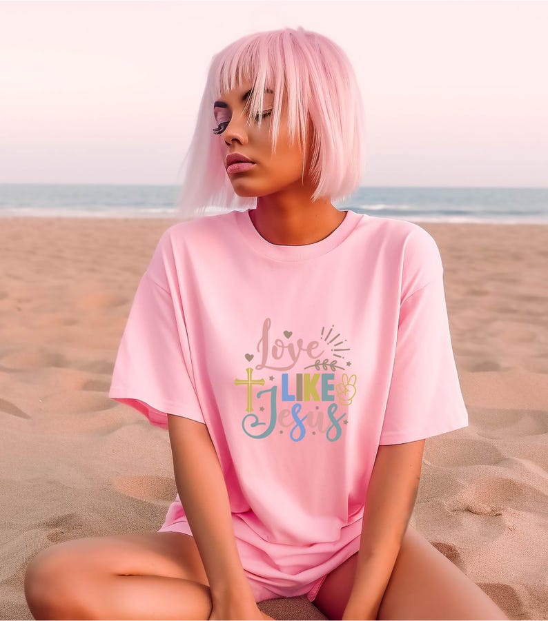 May include: A pink t-shirt with the text "Love Like Jesus" in colorful letters. The shirt features a cross, a peace sign, and hearts.