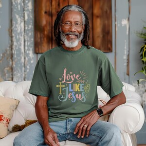 May include: A man wearing a dark green t-shirt with the text "Love Like Jesus" in colorful letters. The shirt features a cross and a peace sign.