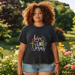 May include: A woman wearing a dark blue t-shirt with the text "Love Like Jesus" in colorful letters. The shirt features a cross, a peace sign, and hearts.