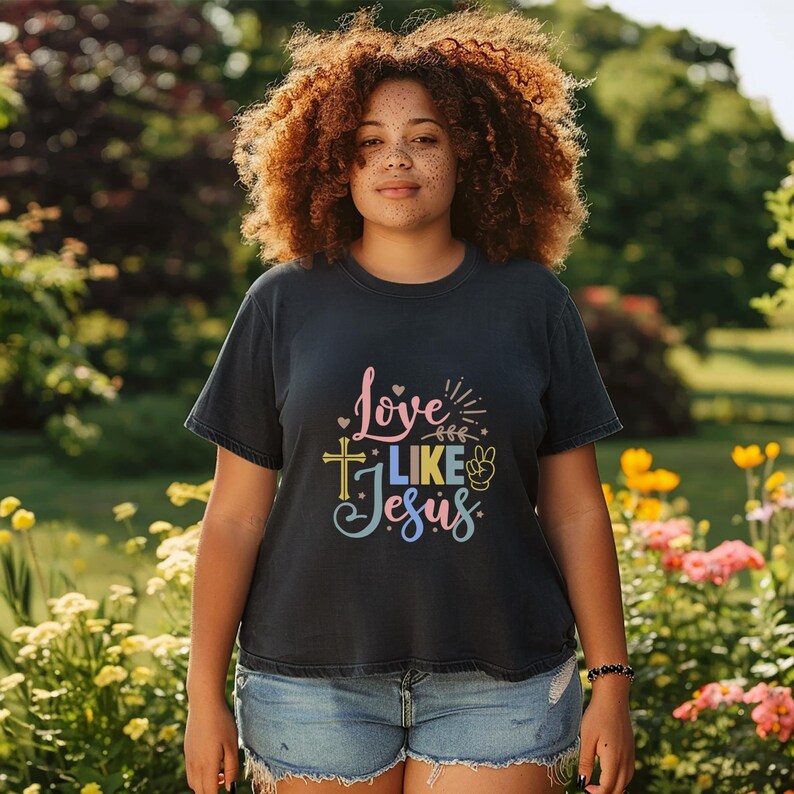 May include: A woman wearing a dark blue t-shirt with the text "Love Like Jesus" in colorful letters. The shirt features a cross, a peace sign, and hearts.