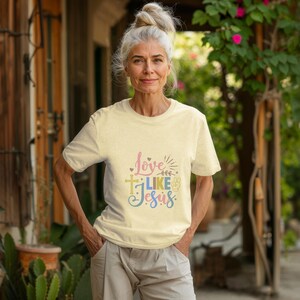 May include: A woman wearing a light beige t-shirt with the text "Love Like Jesus" in colorful letters. The text includes a cross, a peace sign, and a floral design.