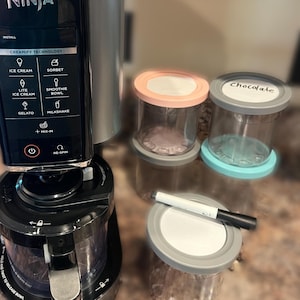 May include: A Ninja CREAMi ice cream maker with a silver and black design. The control panel features options for ice cream, sorbet, and gelato. Includes several clear containers with lids, one labeled "Chocolate". A white marker is also present.