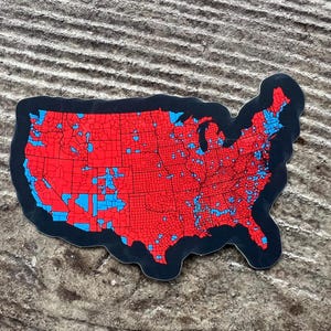 May include: A sticker in the shape of the United States, with a black border. The map is red with blue areas, representing a political map. The sticker is on a textured, gray surface.