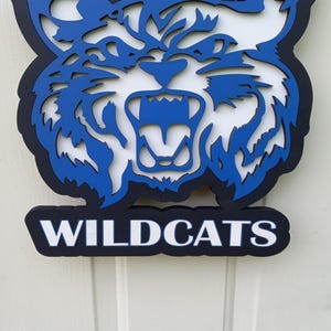 May include: A blue and white wooden Wildcats sign with a cutout of a snarling wildcat head. The sign is hanging on a white door.
