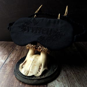 Può includere: Maschera per dormire nera con la parola "WITCH" ricamata in un motivo floreale. La maschera è esposta su un supporto decorativo con una base a forma di teschio e uno sfondo scuro. La maschera per dormire ha un elastico.