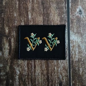 May include: Two embroidered patches on a black background. Each patch features a gold-coloured letter "V" intertwined with green leaves and small white flowers. The patches are rectangular and have a black border.