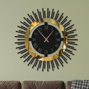 Op de afbeelding: Een zwarte en gouden wandklok met Romeinse cijfers en een zonnestralenontwerp. De klok hangt aan een groene muur boven een bruine bank.